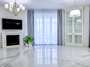 Bright living room with a wall-mounted TV above a white fireplace, a glossy marble floor, and a green potted plant by large windows with sheer curtains.